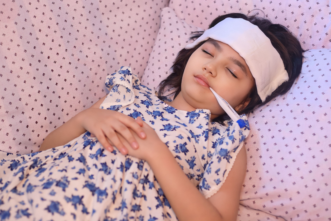 Anak perempuan sedang istirahat di tempat tidur dengan kompres dahi dan termometer, menandakan kondisi demam.