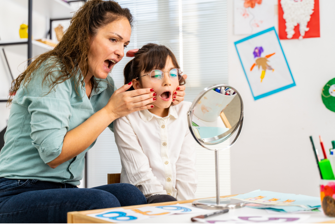Seorang terapis atau orang dewasa membantu anak berlatih bicara di depan cermin. Anak memakai kacamata dan membuka mulut untuk meniru gerakan mulut yang dicontohkan orang dewasa.